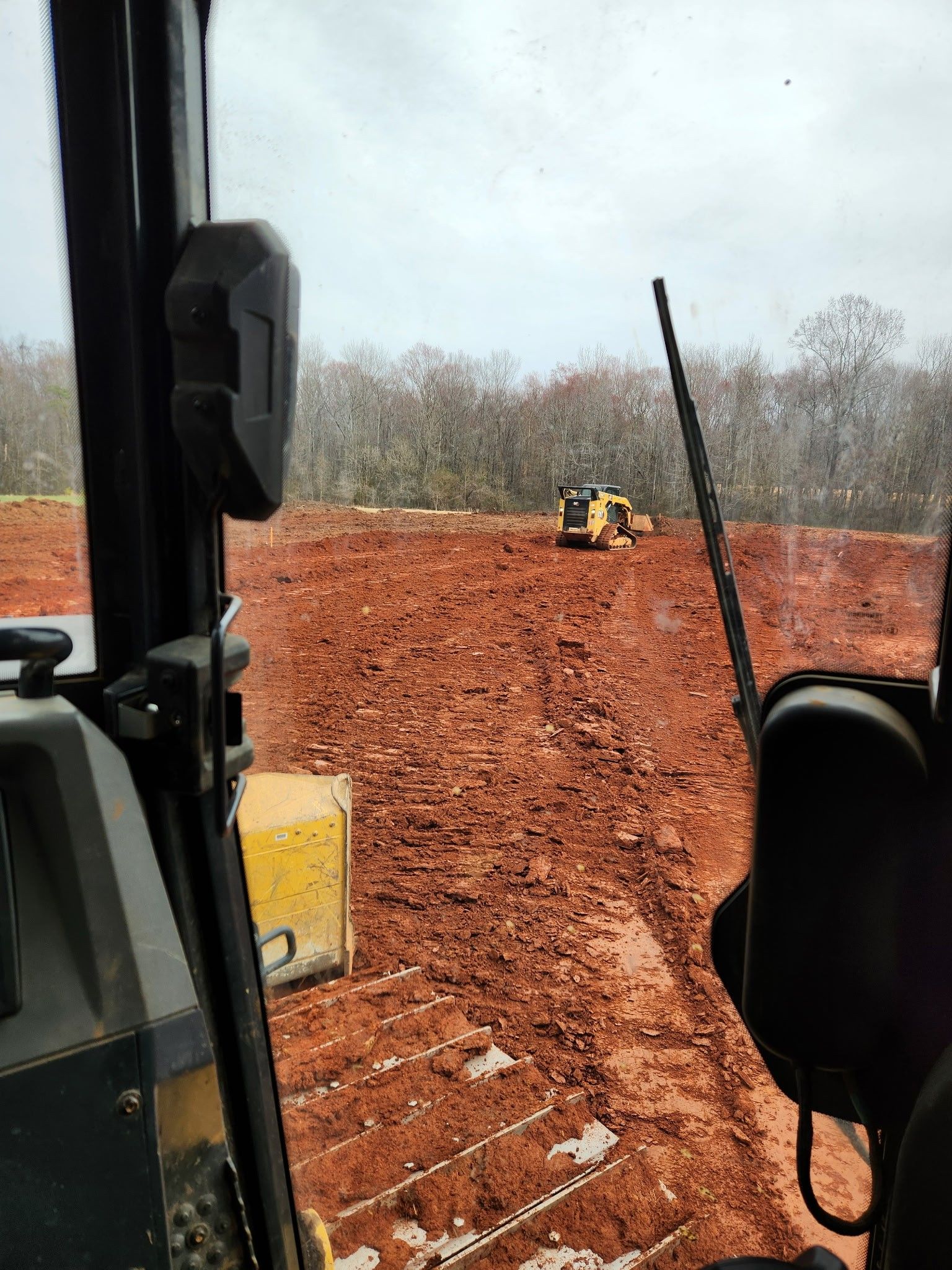 View from an excavator cab of a red dirt construction site with another excavator in the distance.