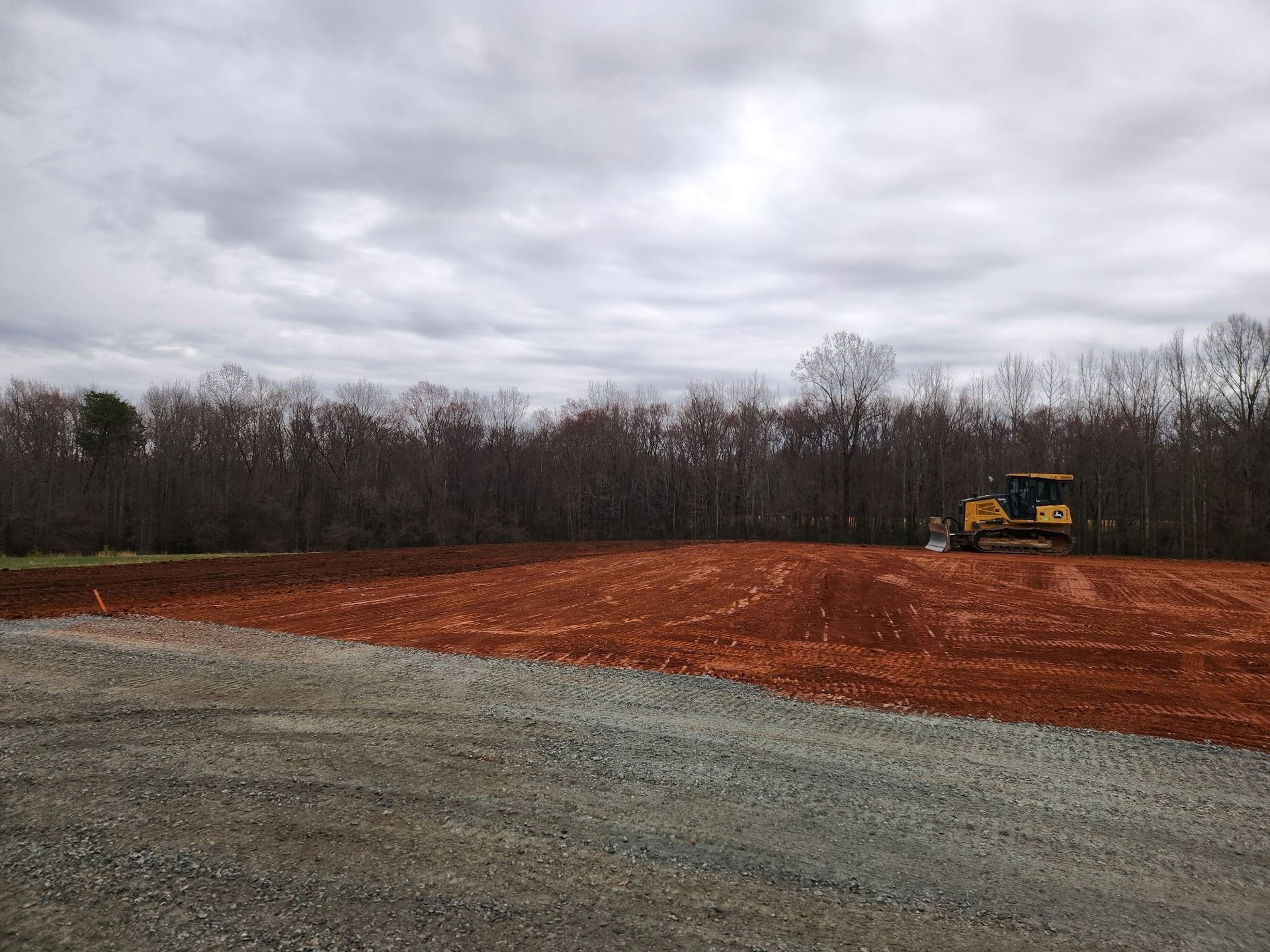 A bulldozer levels red soil on a plot of land, with a line of trees in the background under a cloudy sky.