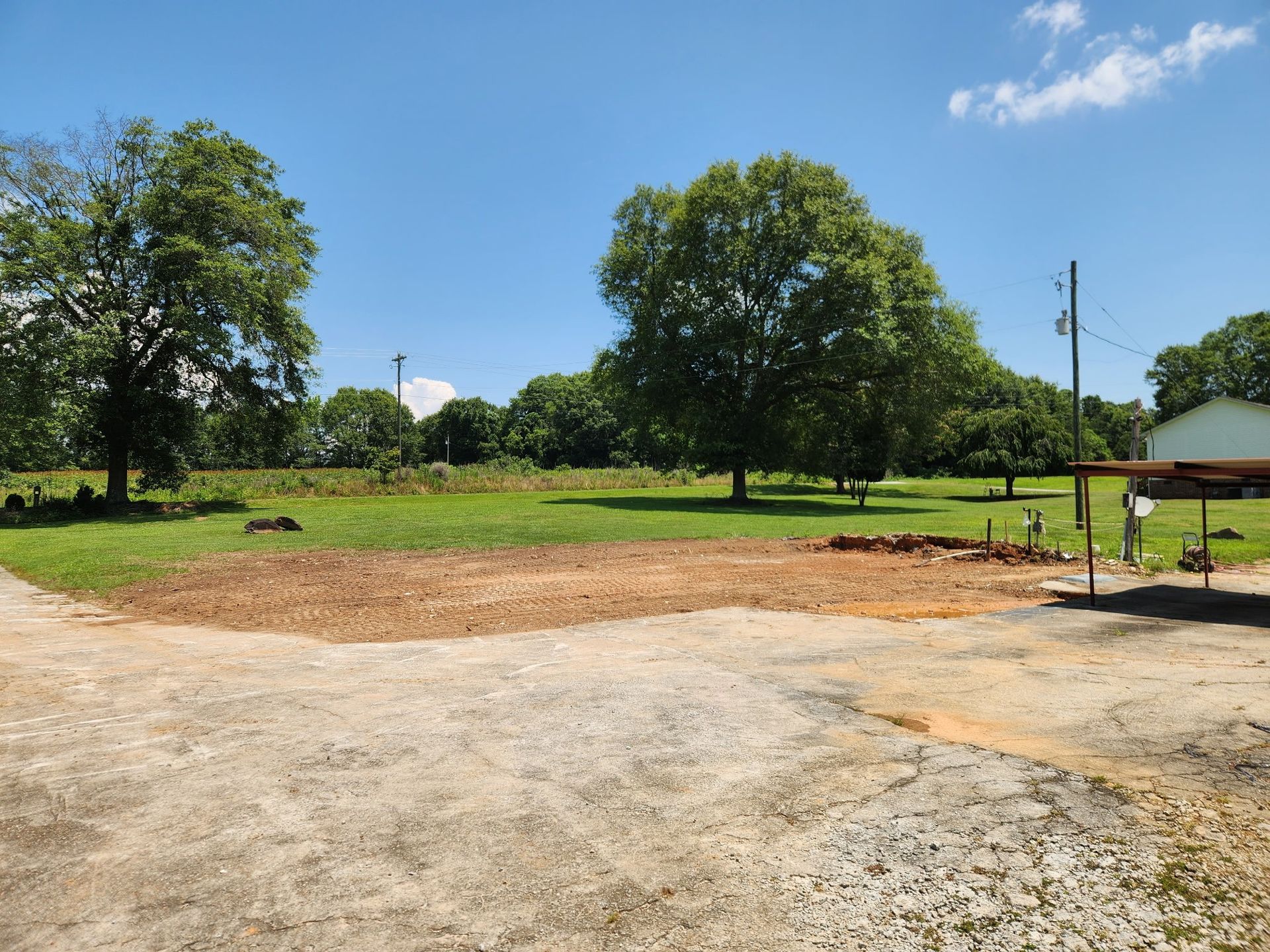 Open, grassy field with gravel and dirt foreground, trees, and a clear blue sky.