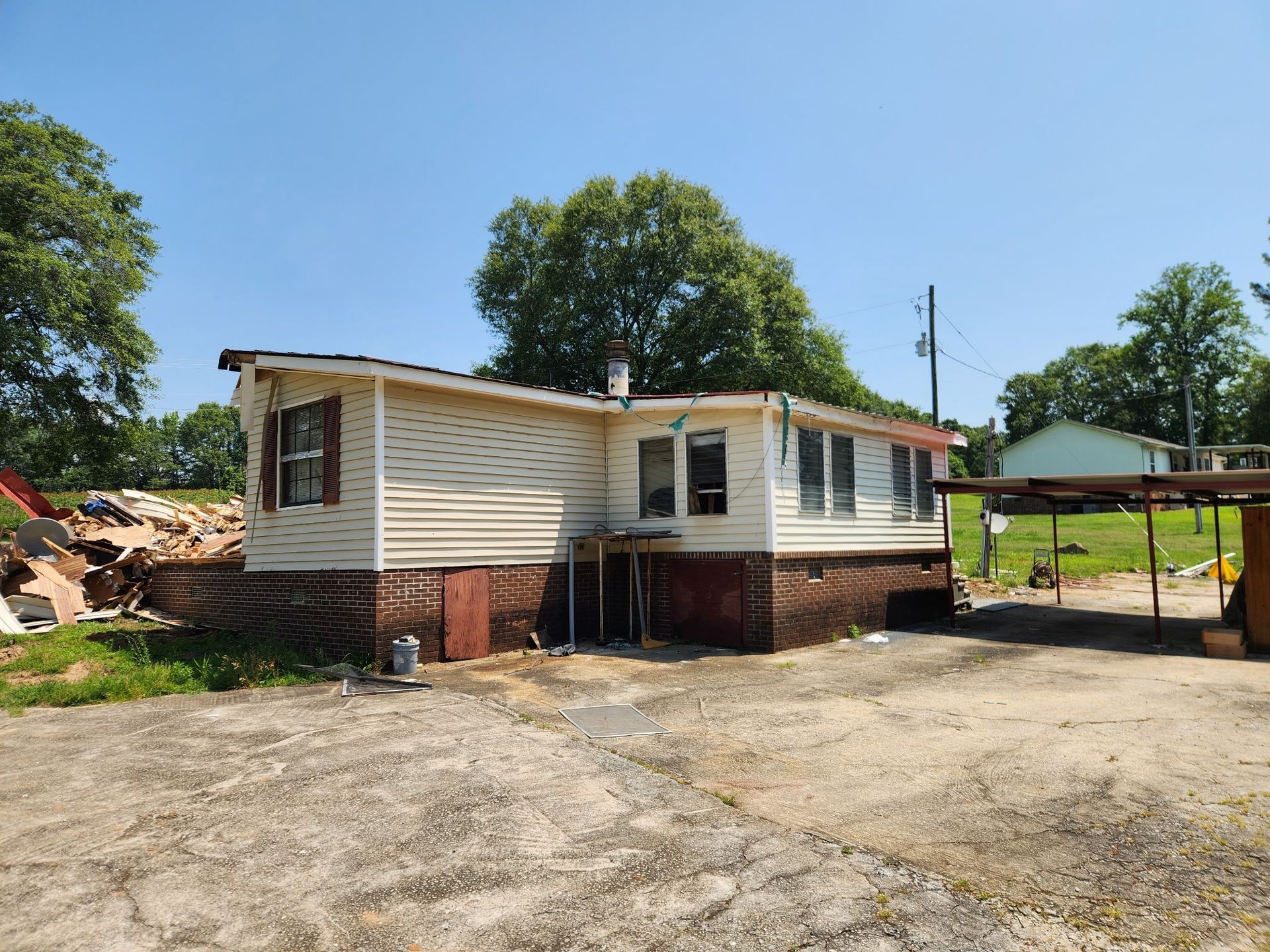Mobile home with beige siding, brown brick foundation, and carport, on a gravel lot under a blue sky.