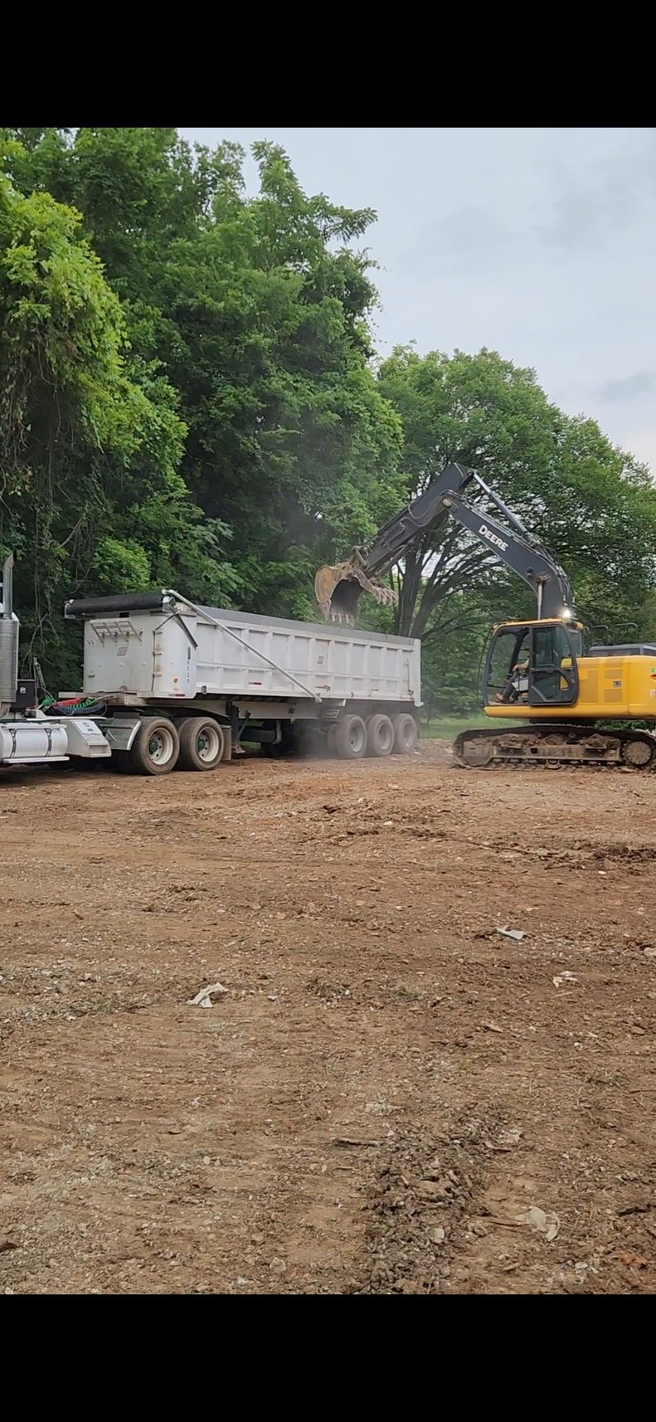 An excavator loading a dump truck with debris on a dirt lot, trees in the background.