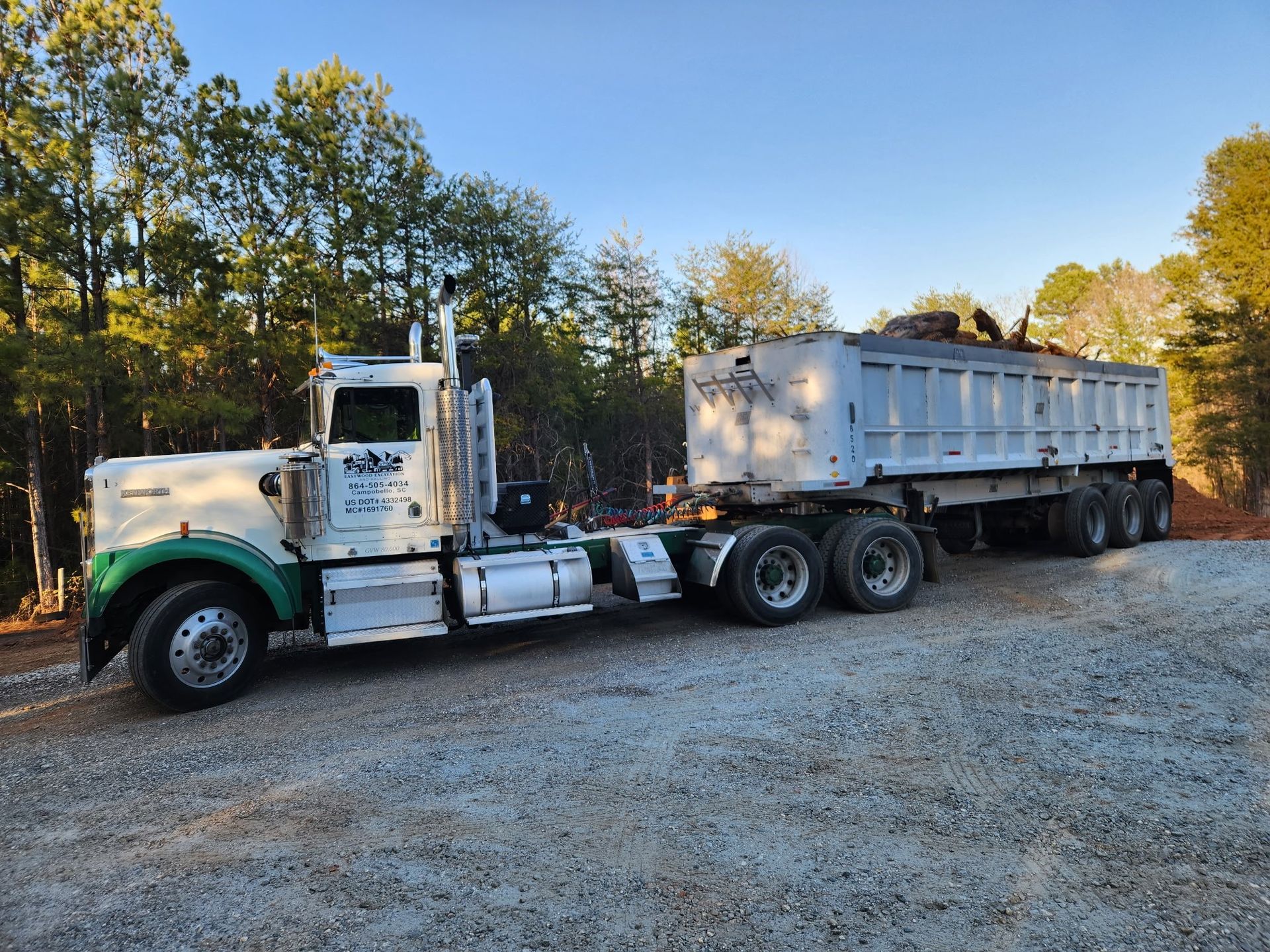 White and green semi-truck with a dump trailer on a gravel road, trees in the background, daytime.