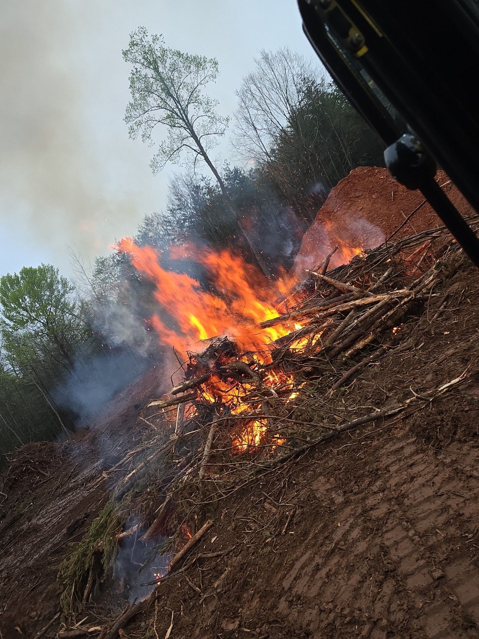 Burning pile of brush and logs on a hillside, flames and smoke billowing.