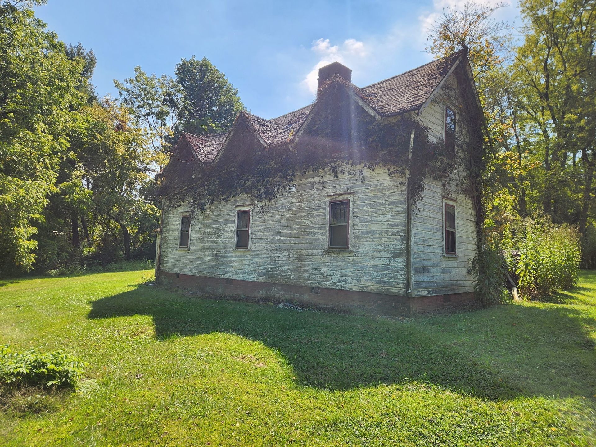 Stone house with ivy and peaked roof, overgrown in a grassy yard, trees in the background, sunny day.