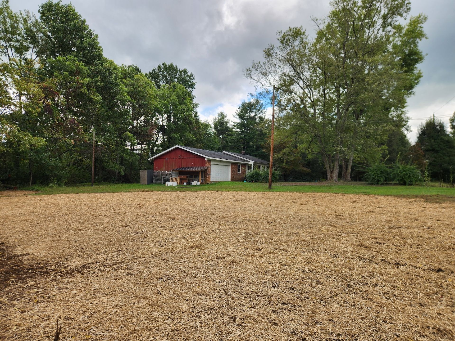A red-roofed building with a garage sits behind a field of wood chips, under a cloudy sky, surrounded by trees.