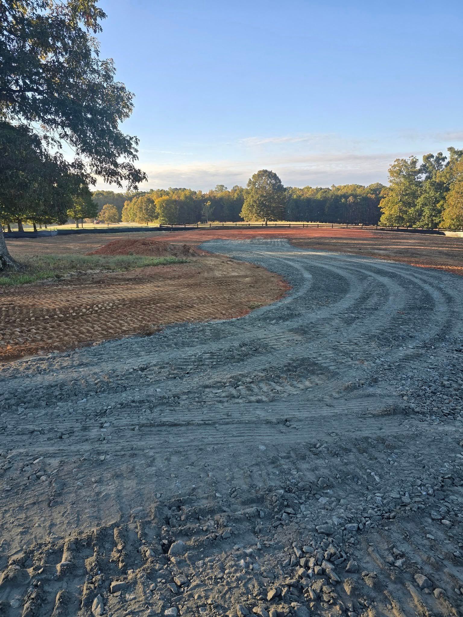 A gravel driveway curves through a construction site, leading to a tree-lined horizon under a blue sky.