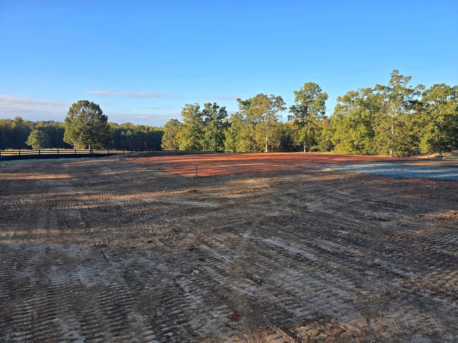 Cleared land, recently graded, with reddish-brown earth and a line of green trees against a blue sky.
