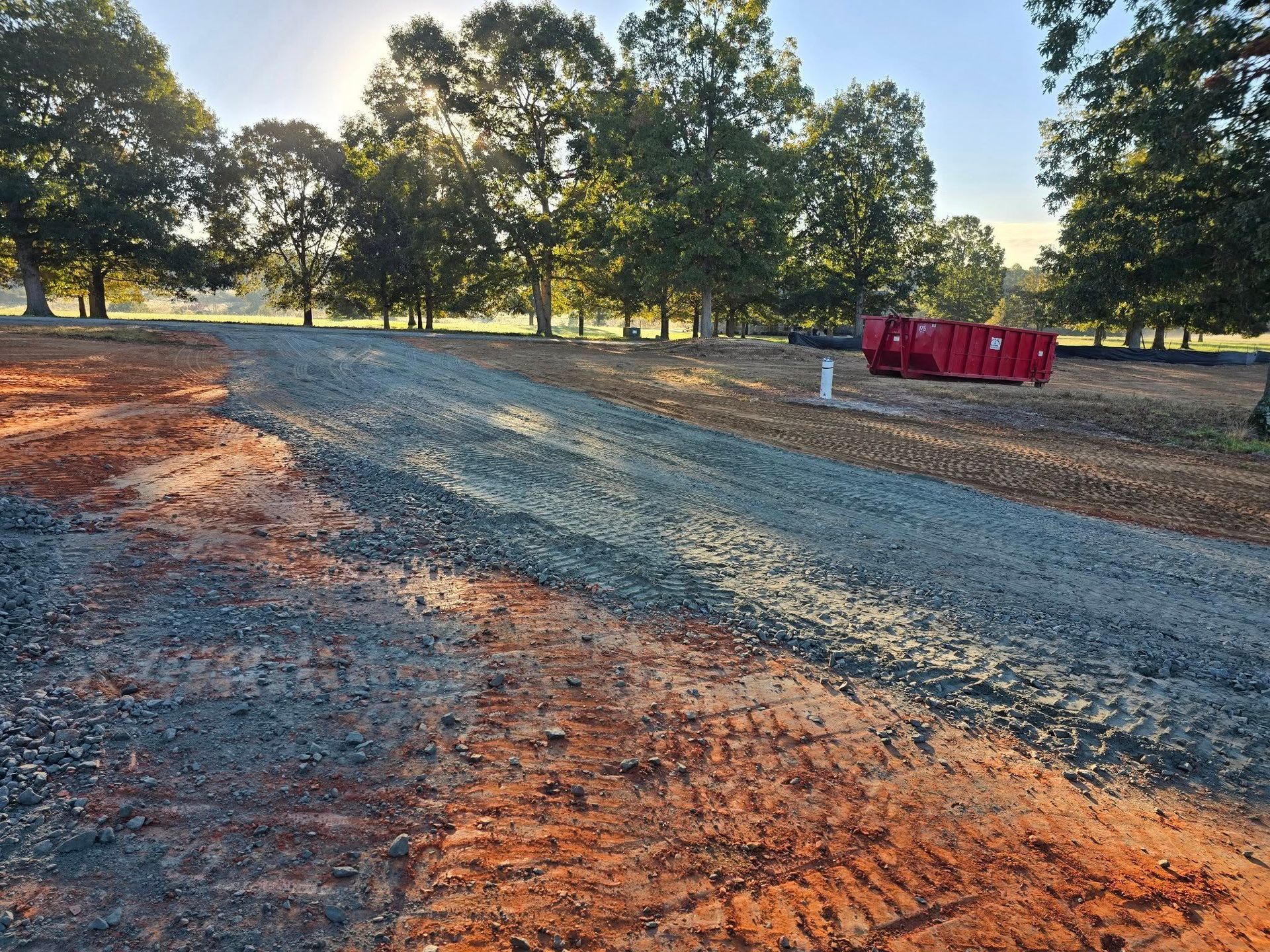 Gravel driveway leading into a grassy field with trees in the background and a red dumpster.