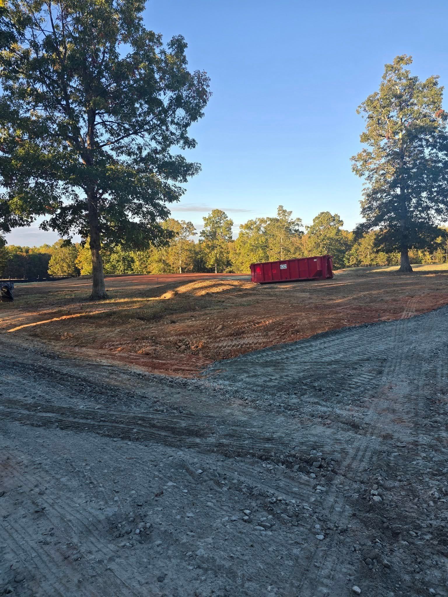 Gravel road leads to grassy field with red container. Trees and blue sky in background.