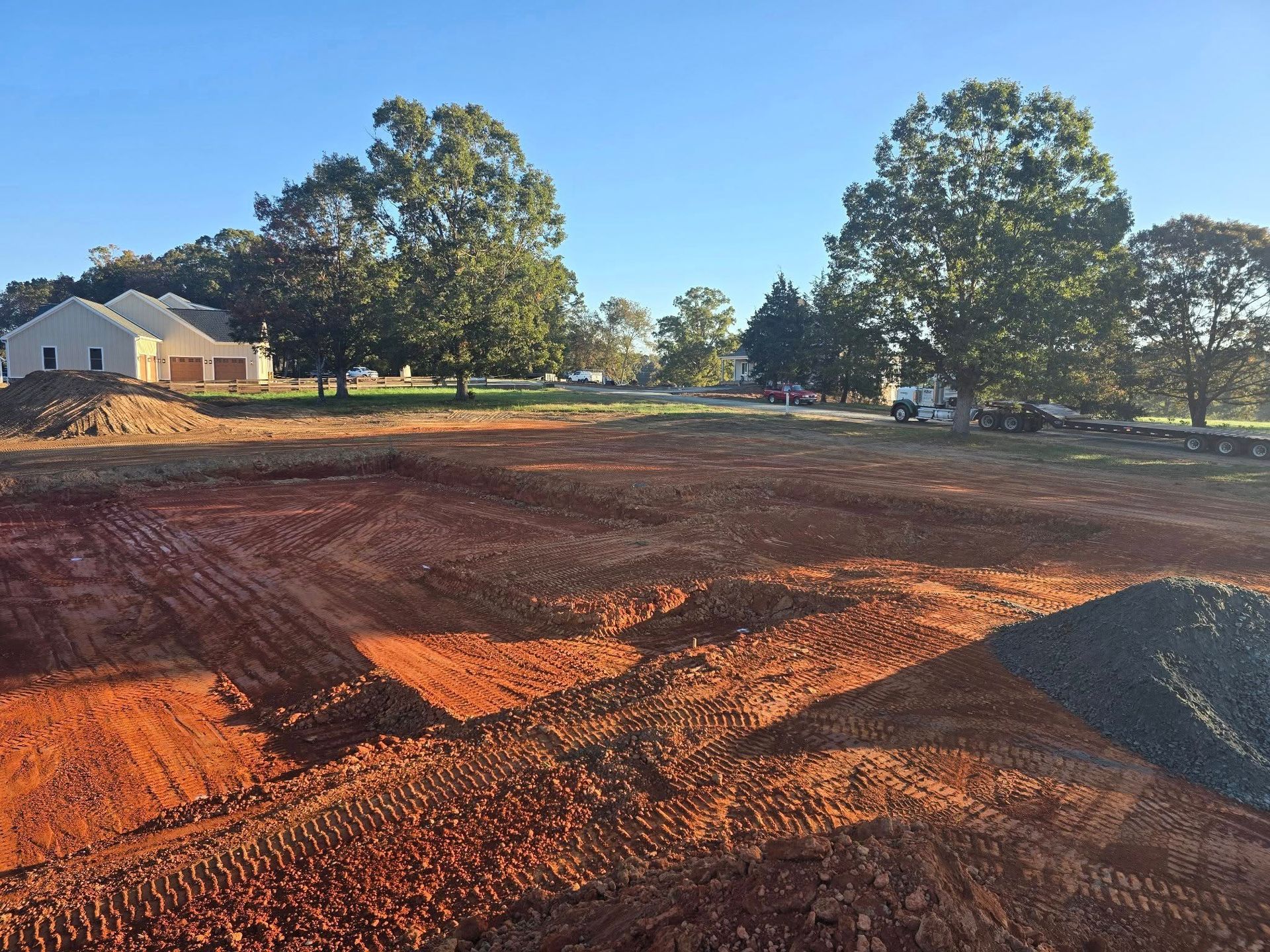Red dirt construction site with two-story house and trees in background.