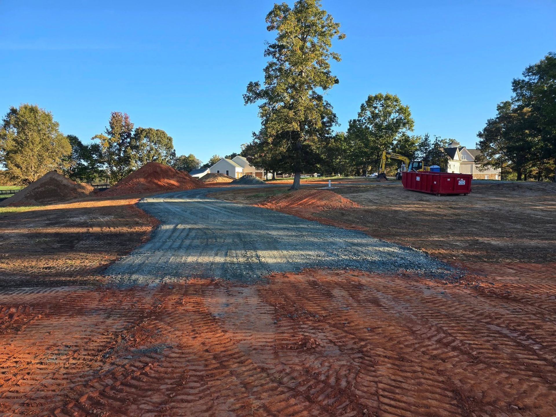 A gravel driveway under construction on a dirt lot, leading toward trees and houses under a blue sky.