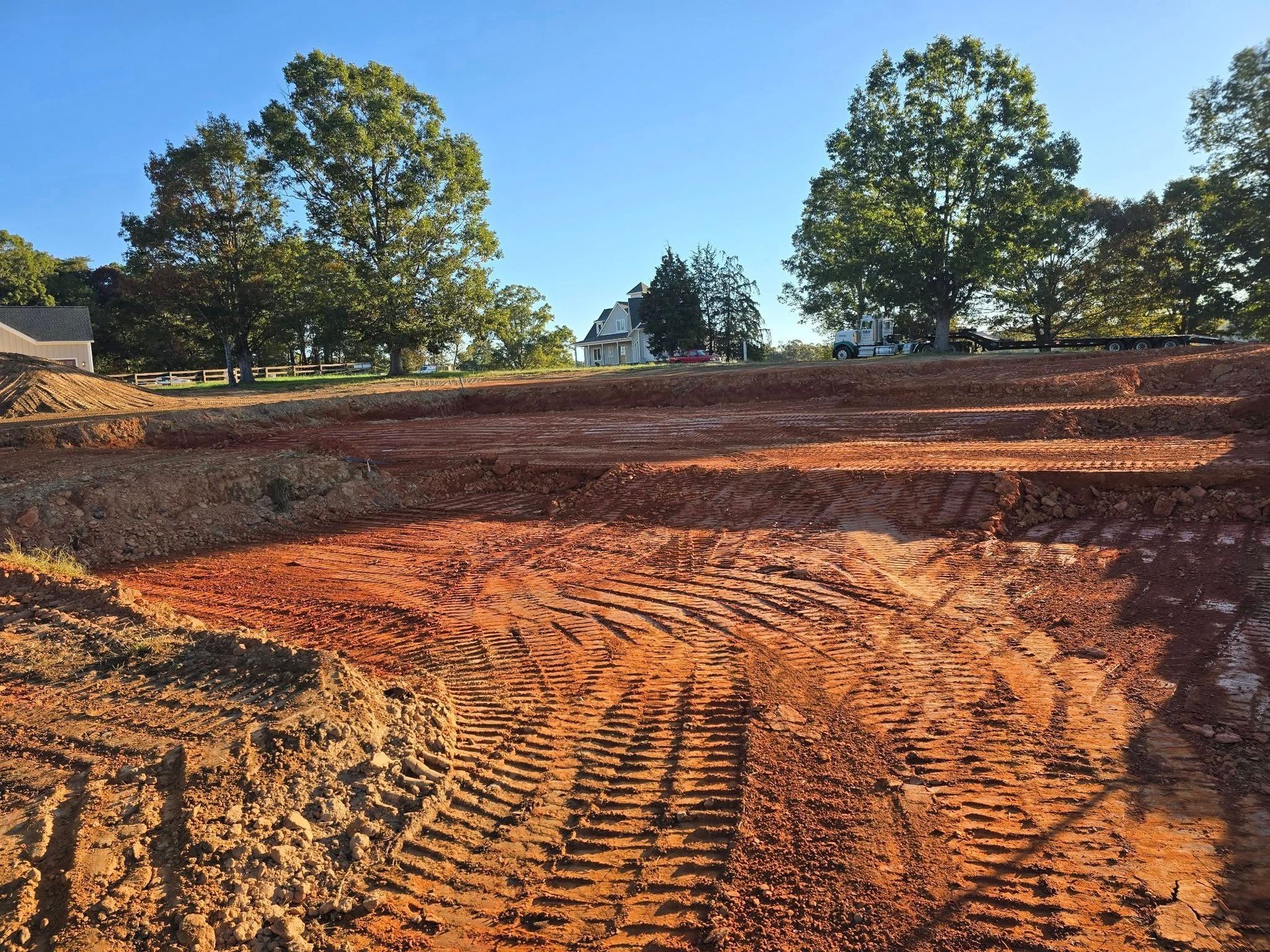 Construction site with red soil, tire tracks, trees, and a house in the background under a blue sky.