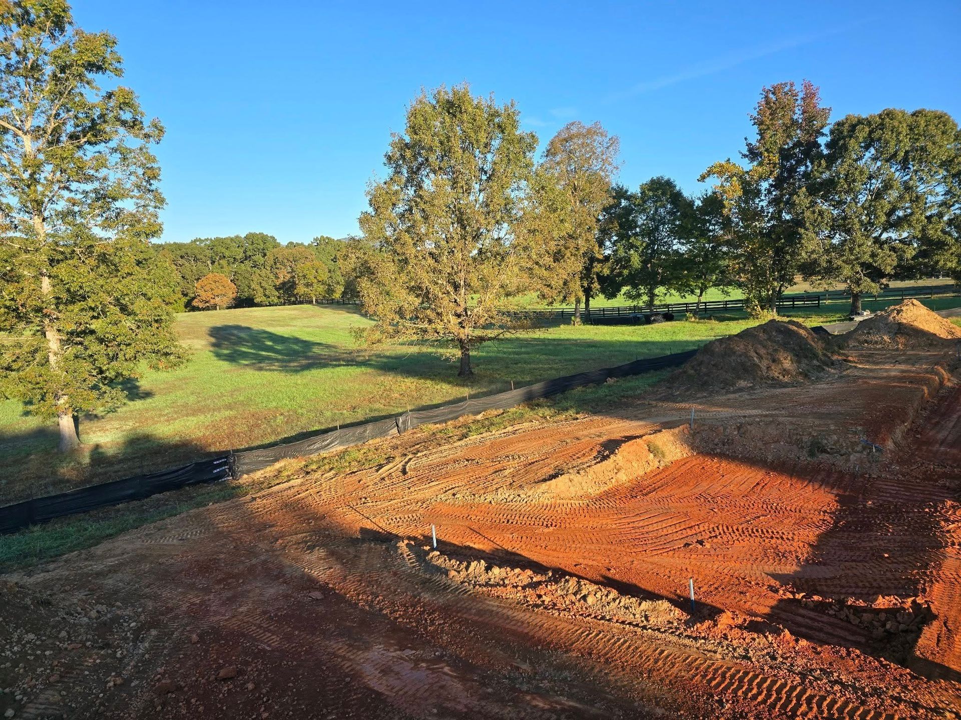 Dirt excavation with surrounding grassy field and trees under a blue sky.