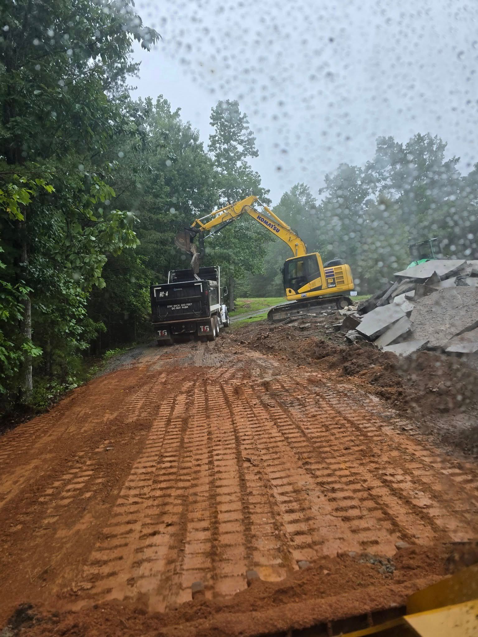 A yellow excavator working with a grapple attachment, moving debris in a muddy clearing near trees.