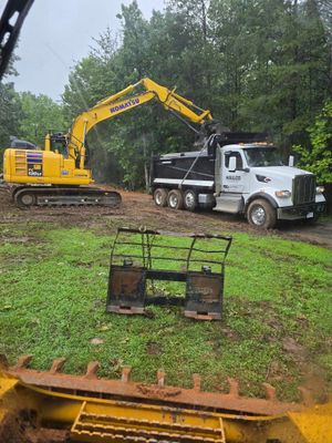 Yellow excavator loading a white dump truck in a muddy, wooded area. A grapple attachment is in the foreground.