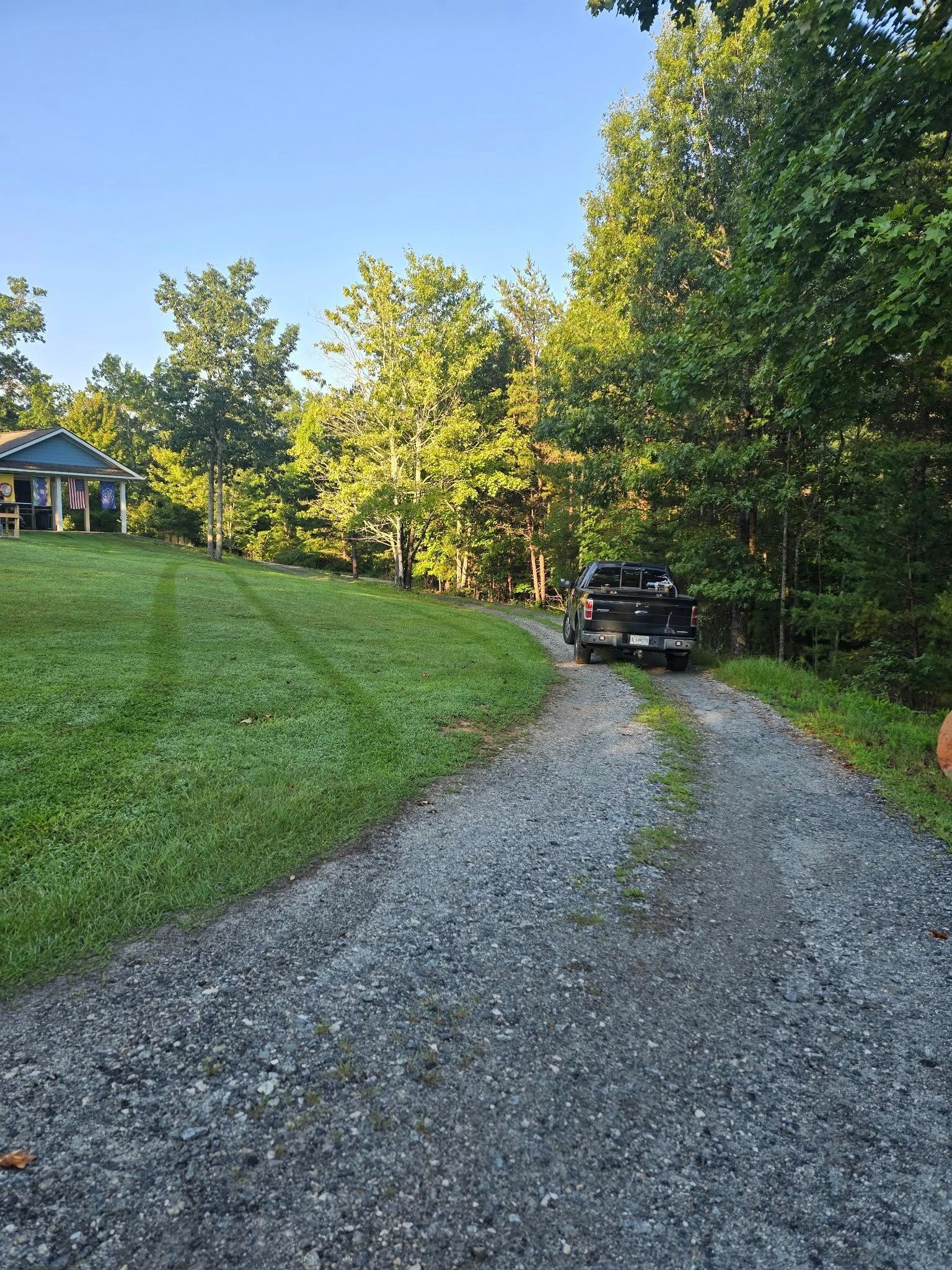 Gravel driveway leading to a truck carrying materials, with a grassy hill and trees; a blue house in the background.