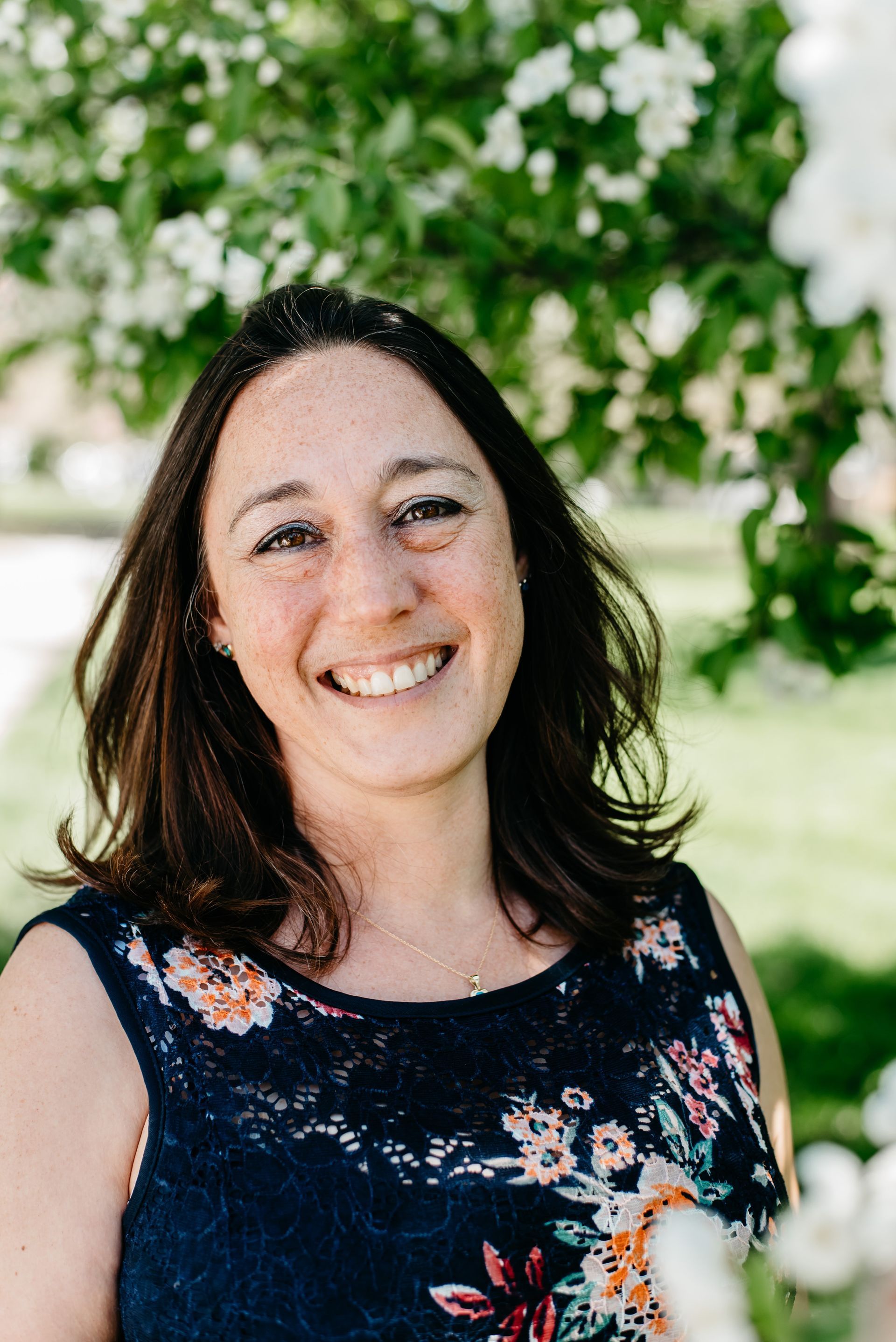 A woman in a floral dress is smiling in front of a tree.