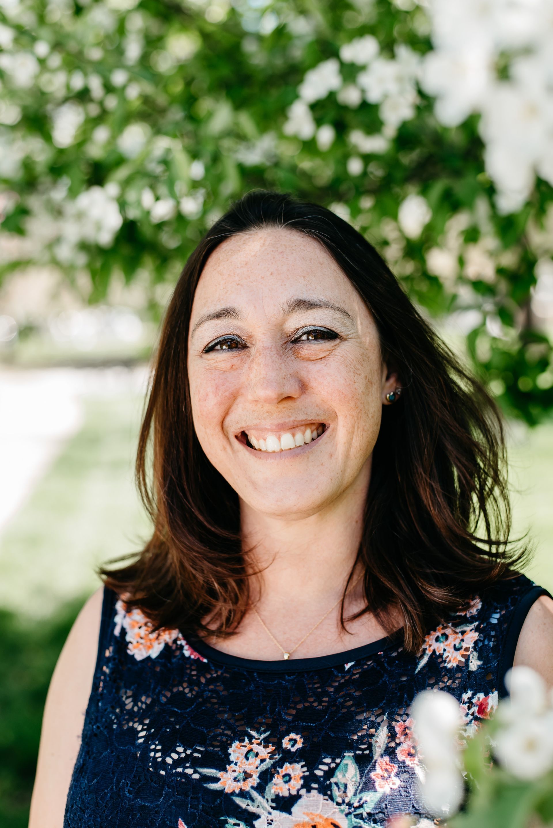 A woman is smiling in front of a tree with white flowers.