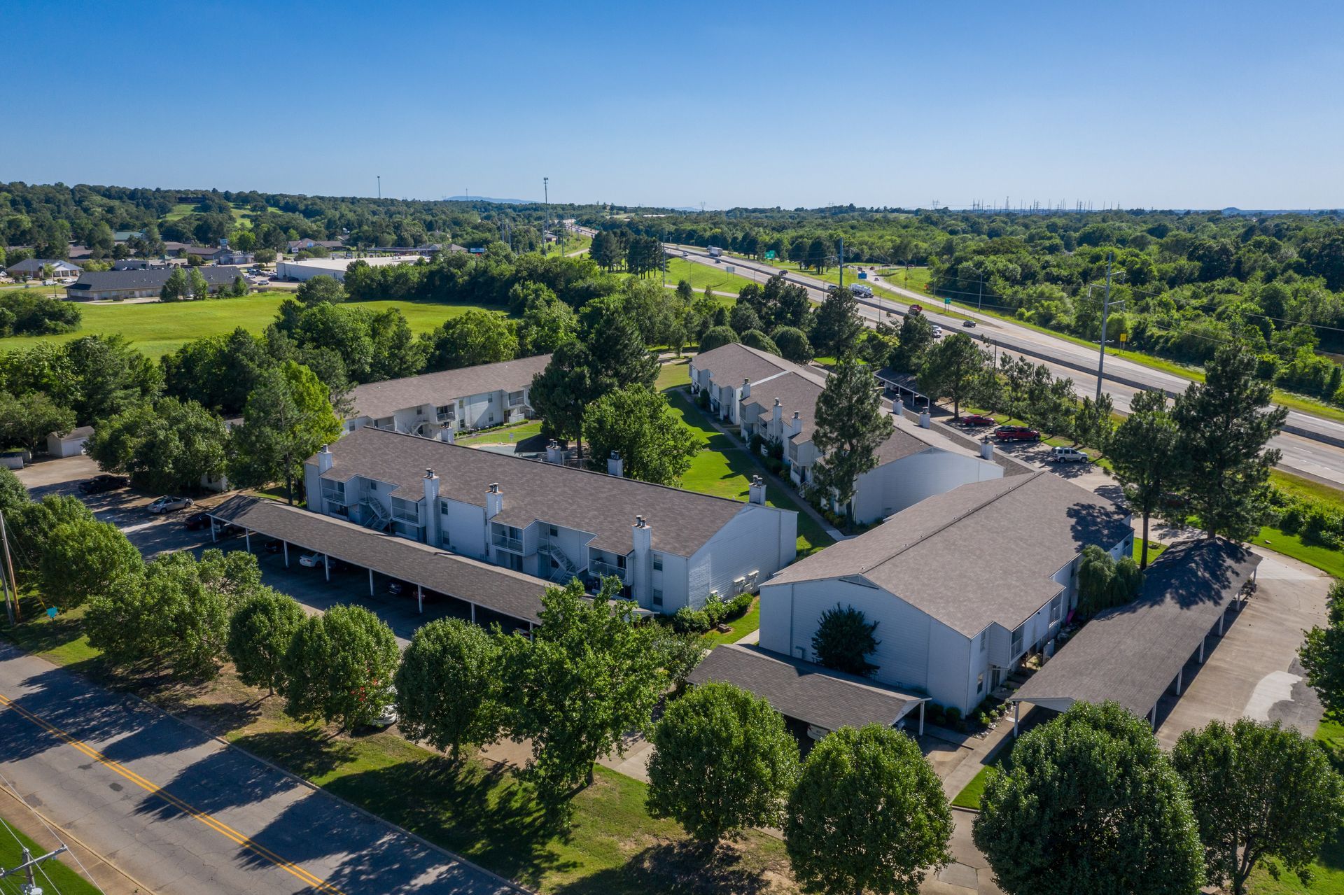 Aerial view of a white apartment complex with attached carports, surrounded by trees and a highway.