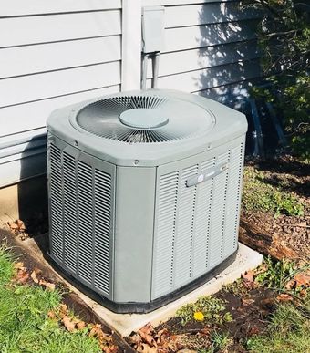 Outdoor central air conditioner unit beside a house, with sunlit siding and a grassy yard.