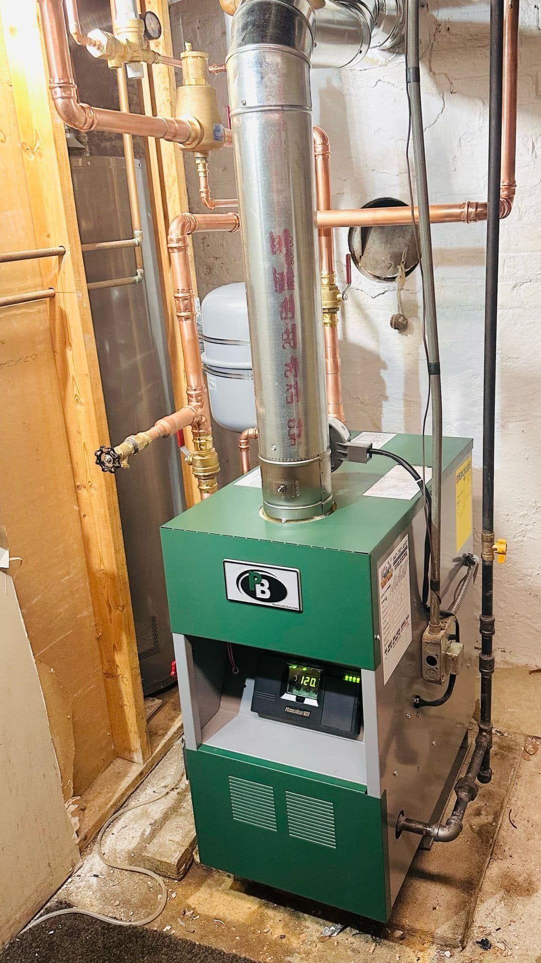 Green boiler unit with metal vent pipes in a utility basement, surrounded by exposed framing and plumbing.