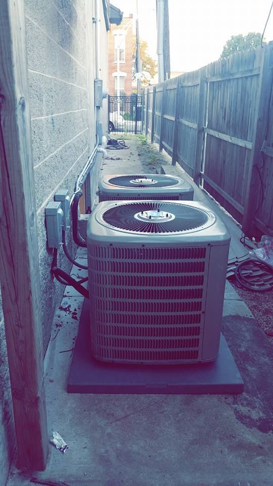 Two outdoor HVAC units in a narrow alley beside a building and fence.