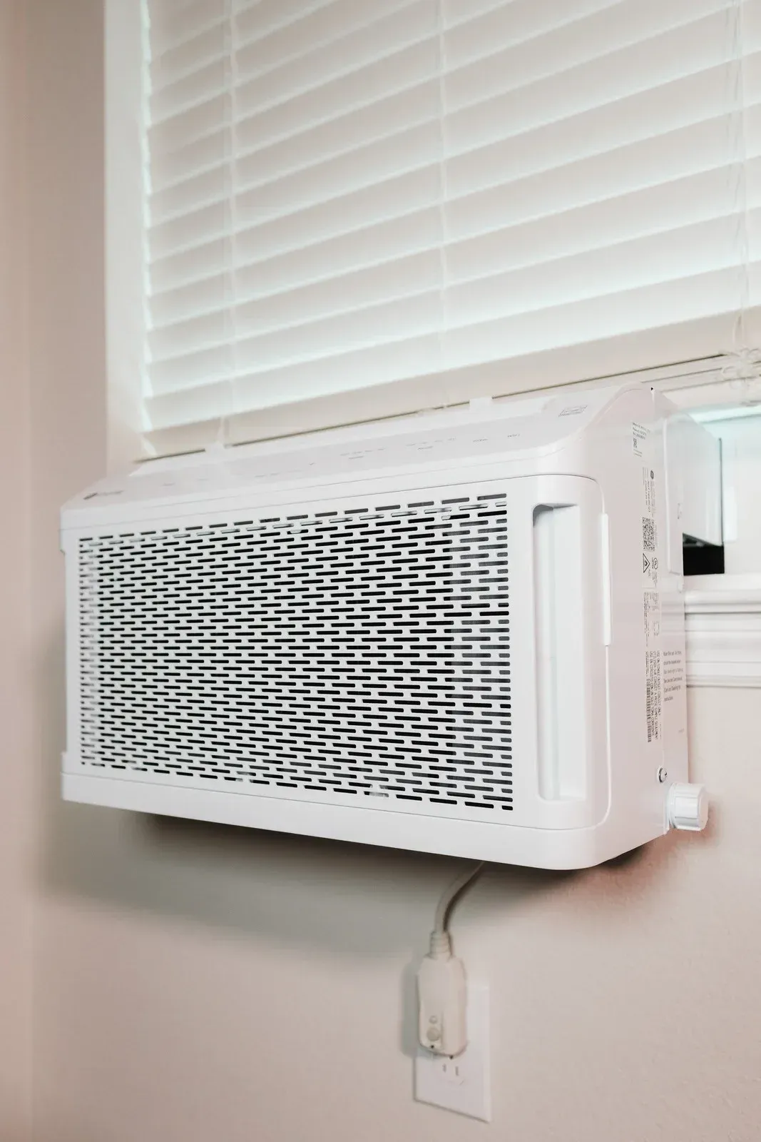 Window air conditioner mounted below a white blind, with cord and outlet on a beige wall