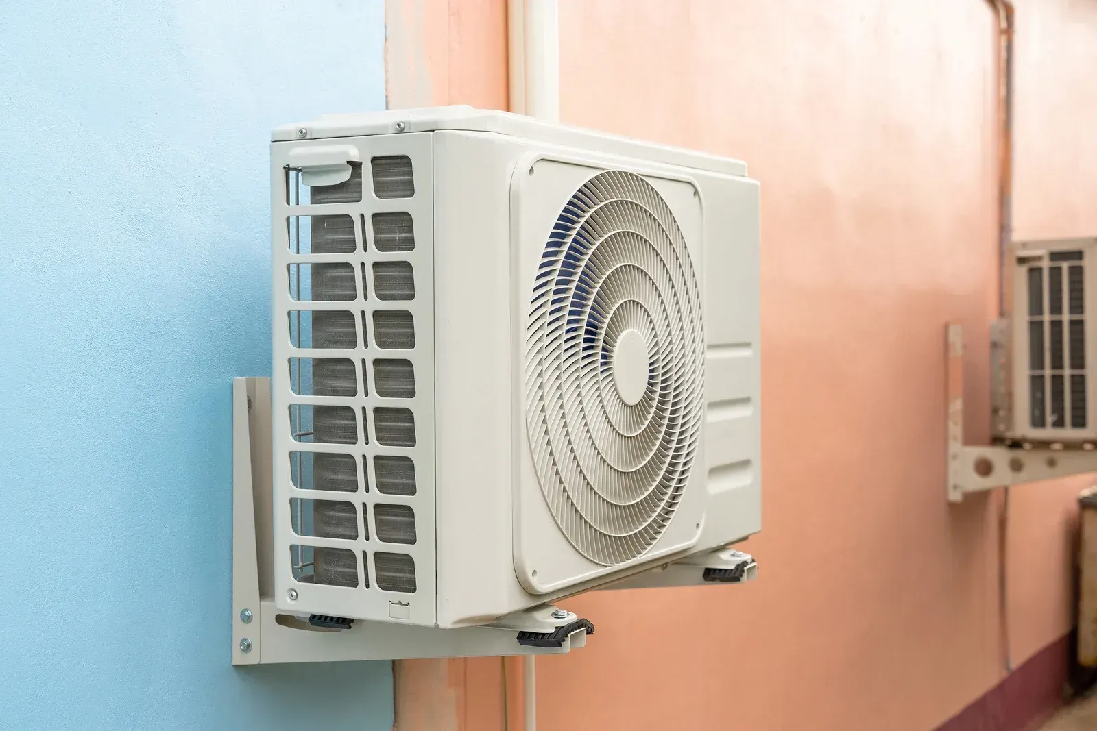 White outdoor air conditioner units mounted on a pastel wall, one close-up with a fan grille visible