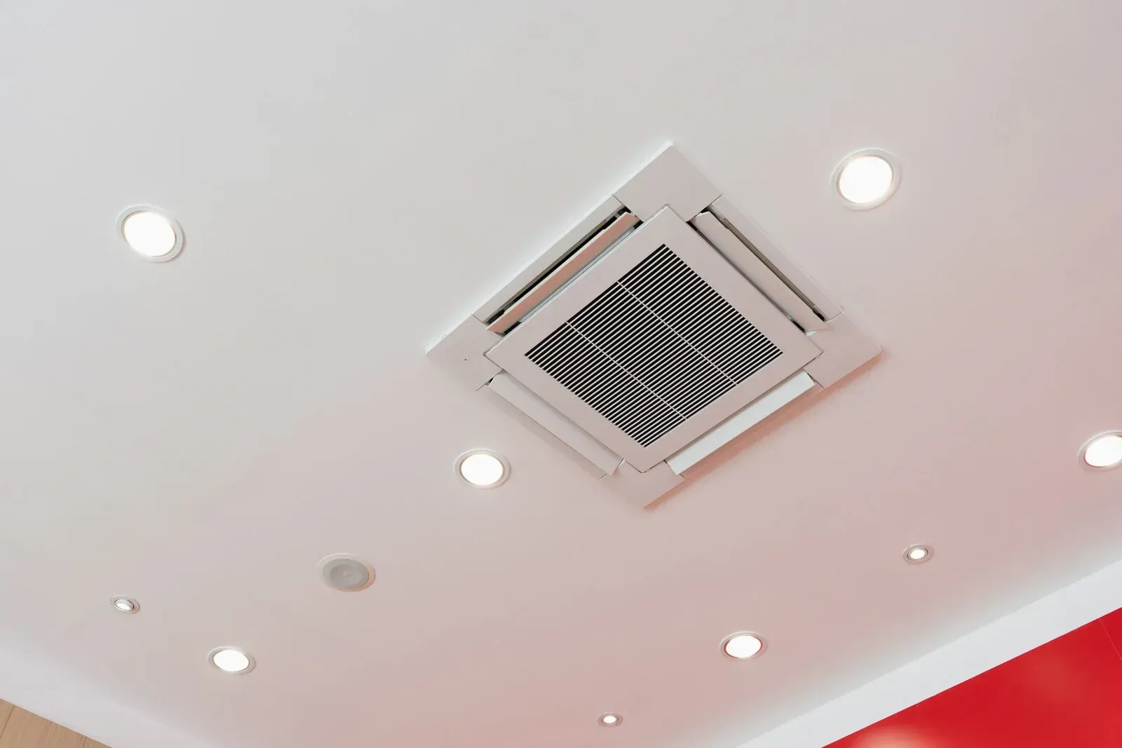 Ceiling with recessed lights and a central air vent in a white room with a red wall edge