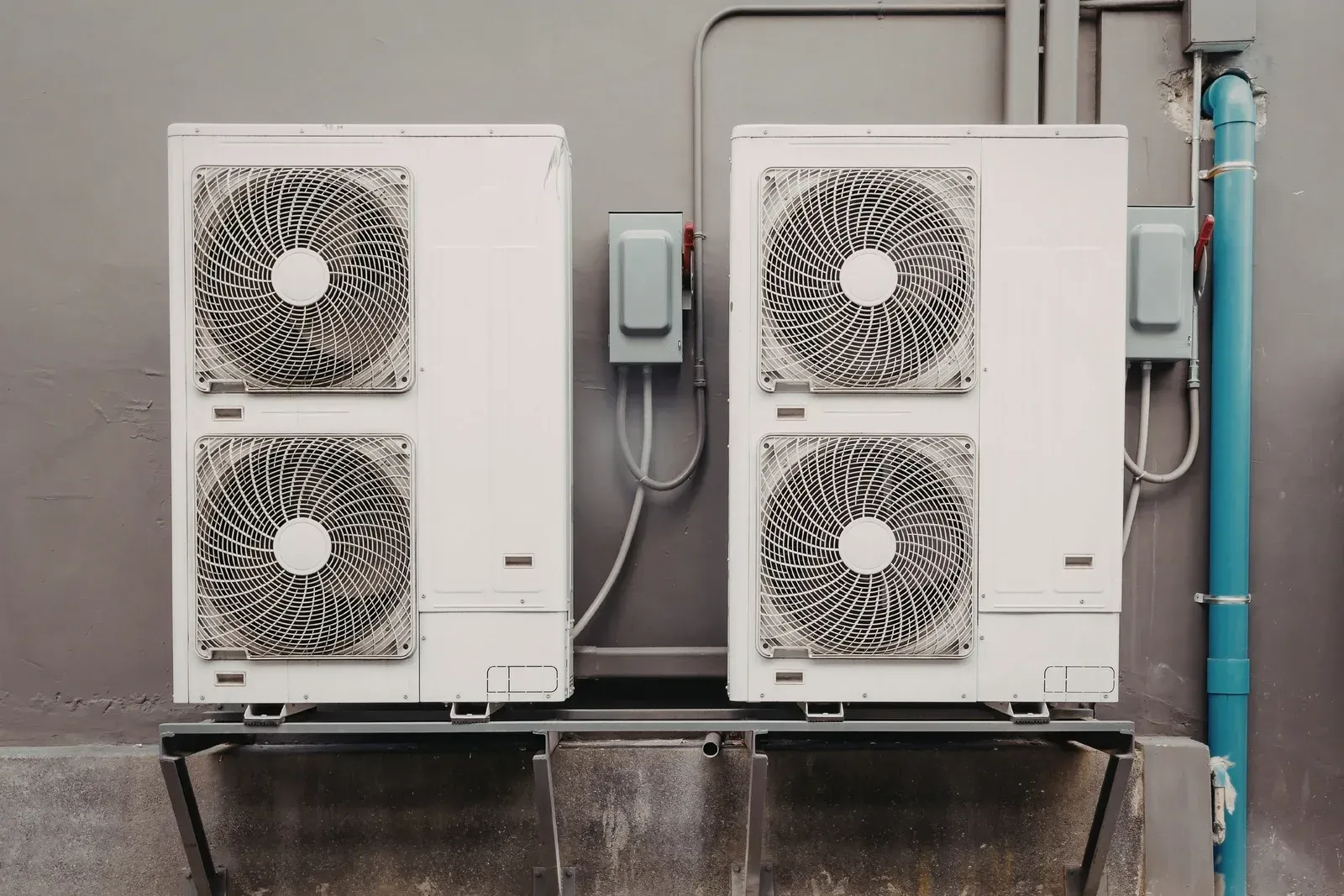 Two outdoor heat pump units mounted on a concrete platform against a gray wall