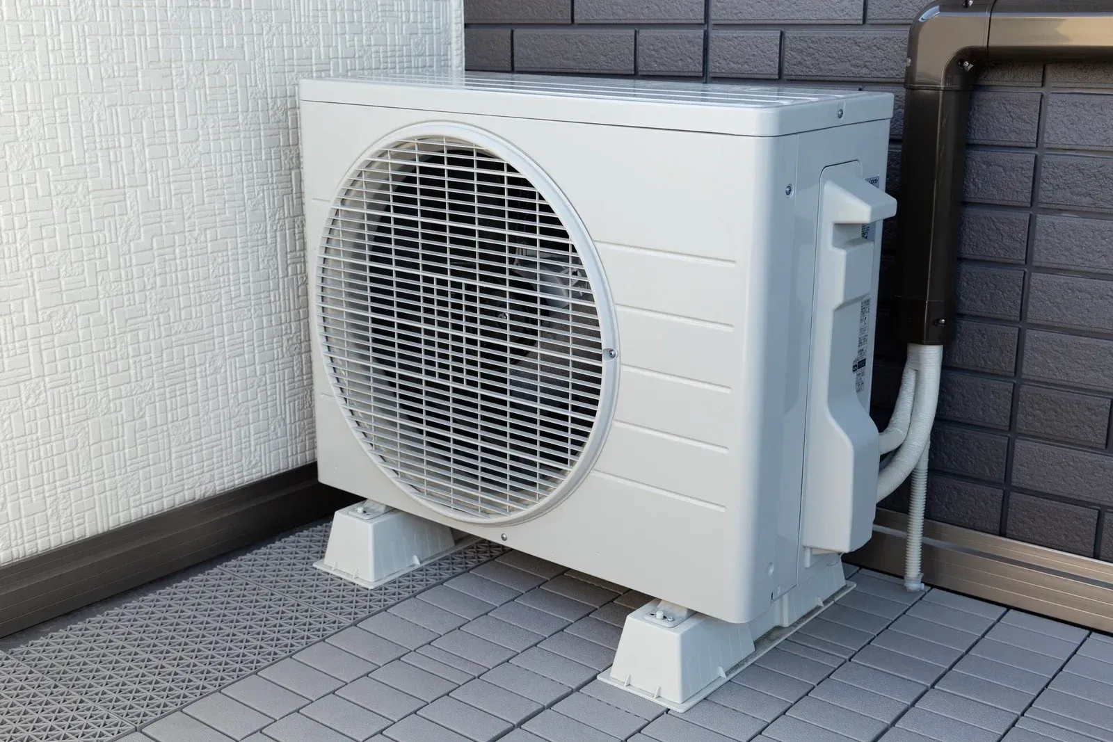 Outdoor white air conditioning unit mounted beside a brick wall on a tiled patio.