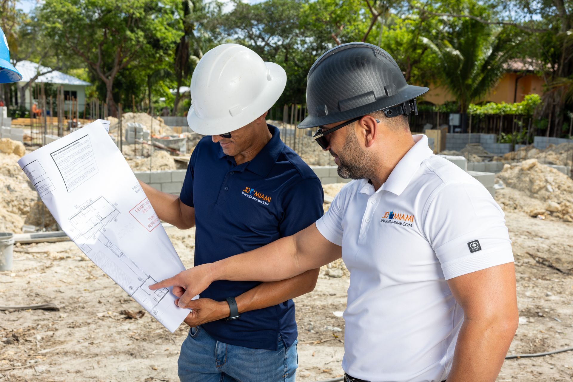 Two men wearing hard hats examine blueprints at a construction site.