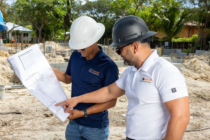 Two men wearing hard hats examine blueprints at a construction site.