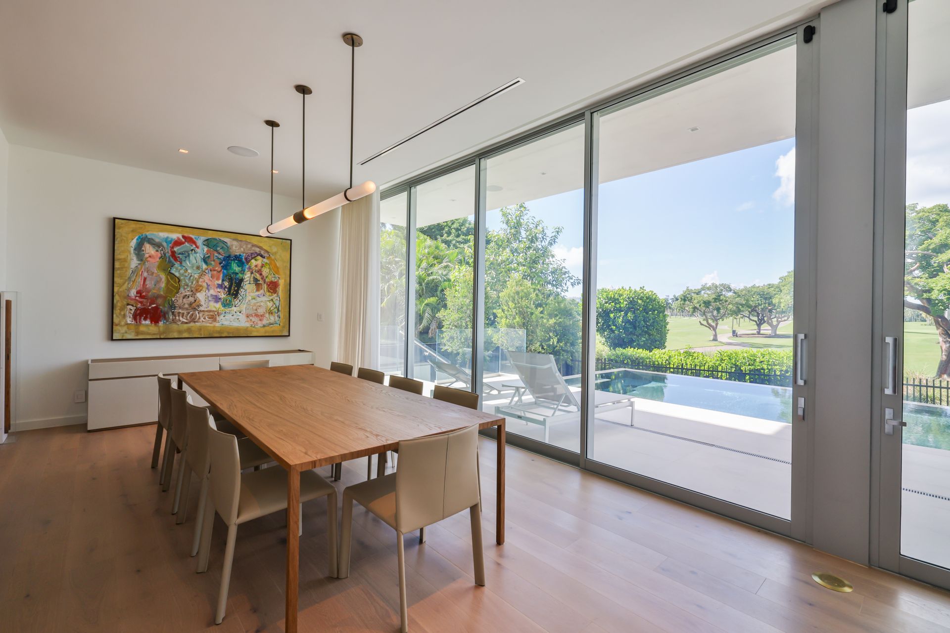 Dining room with a long wooden table, chairs, and large windows overlooking a pool and trees.