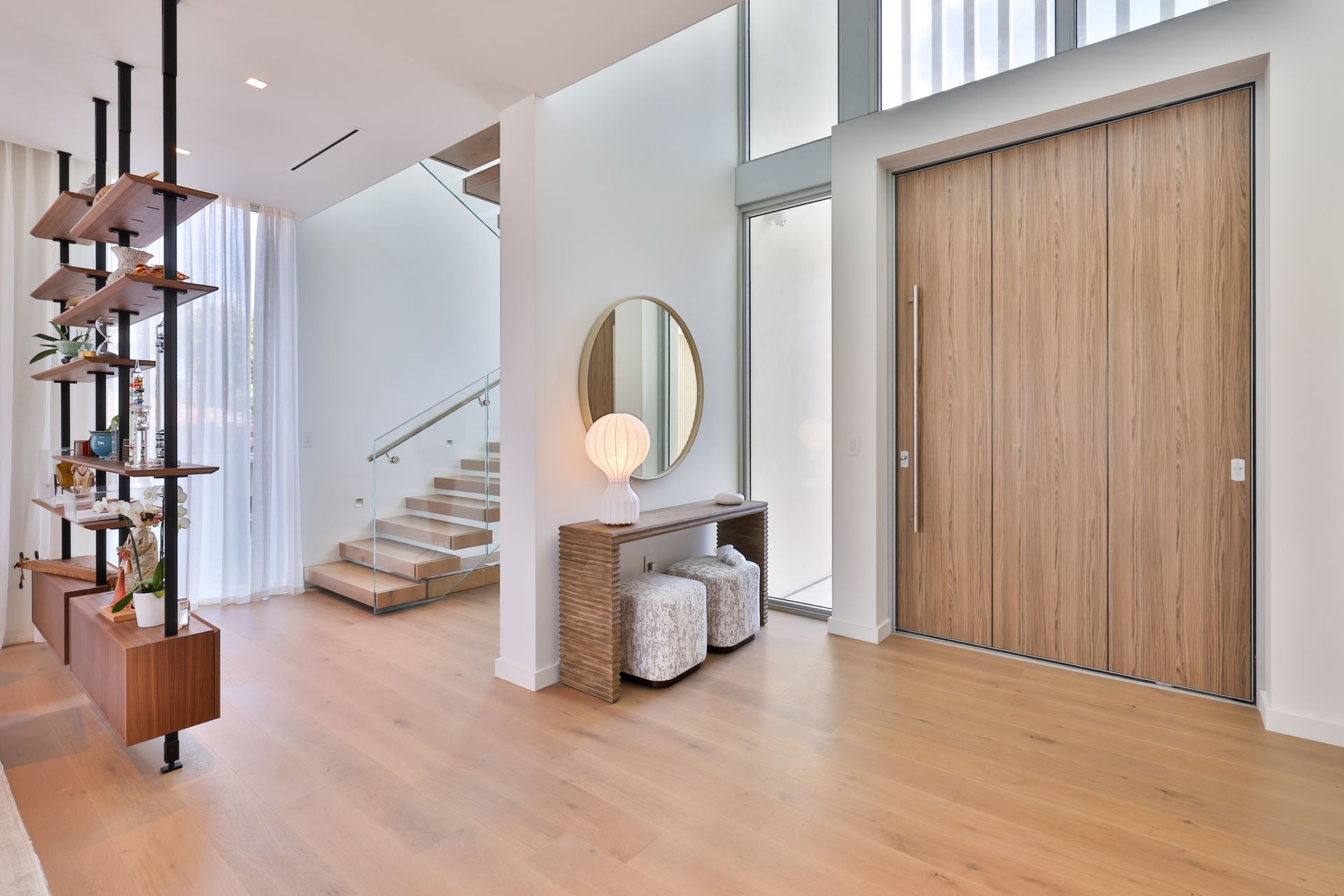 Modern entryway with wooden door, stairs, console, and shelving unit. Light wood flooring and white walls.
