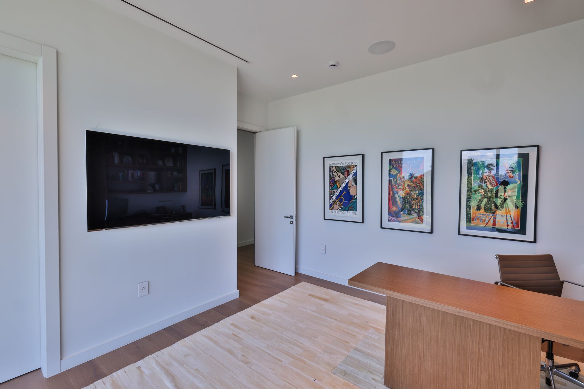 Office with white walls, TV, artwork, desk, and area rug. Wooden floor, door.