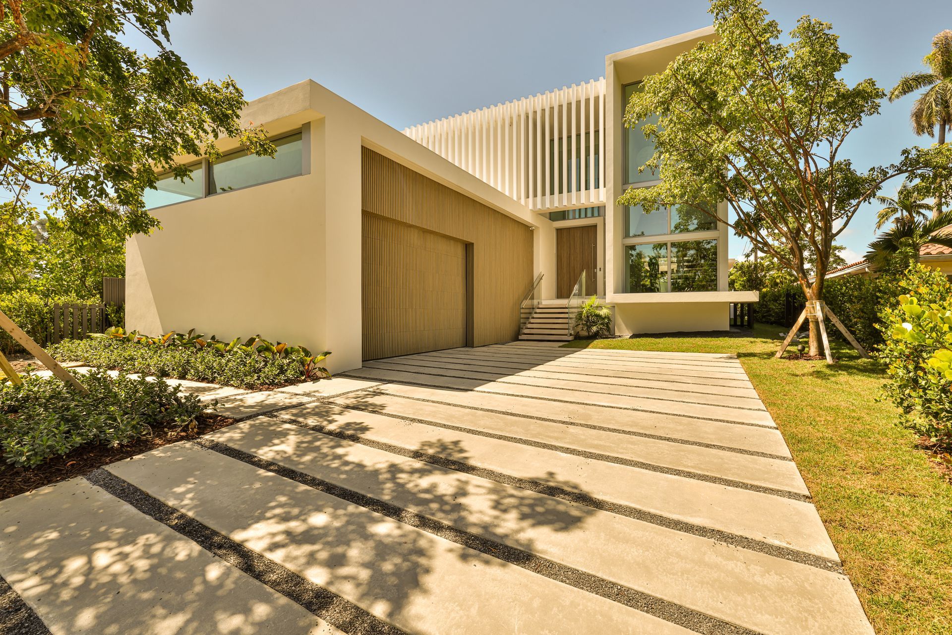 Modern white house with stone driveway and wooden garage door. Lush greenery surrounds the house on a sunny day.