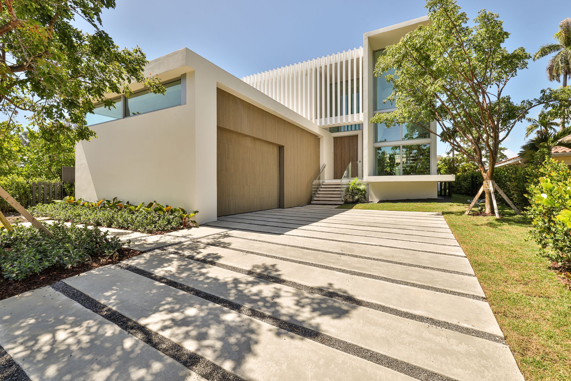 Modern white house with stone driveway and garage, surrounded by trees and grass, under a blue sky.