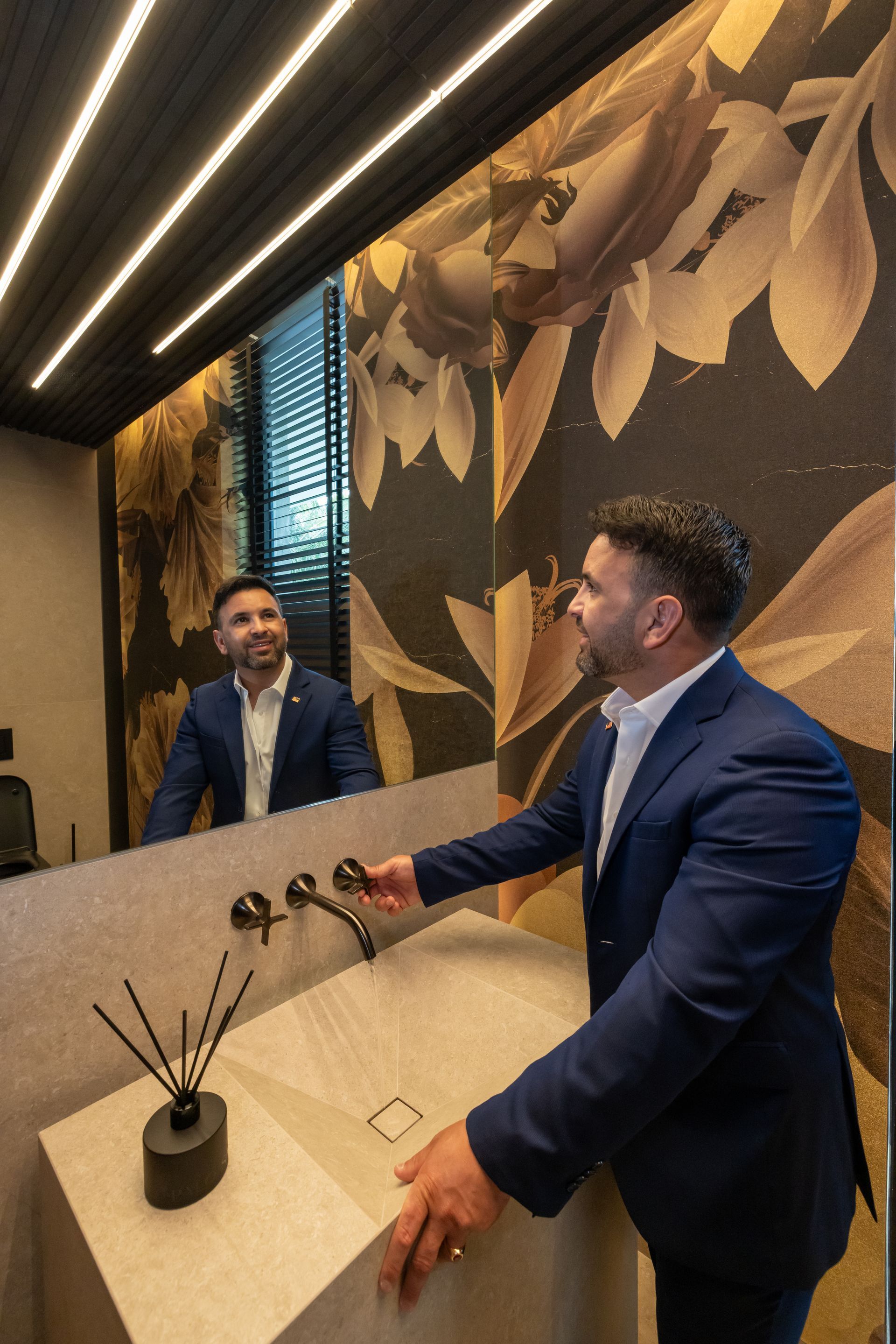 Man in suit at sink, looking in mirror. Decorative floral wall.