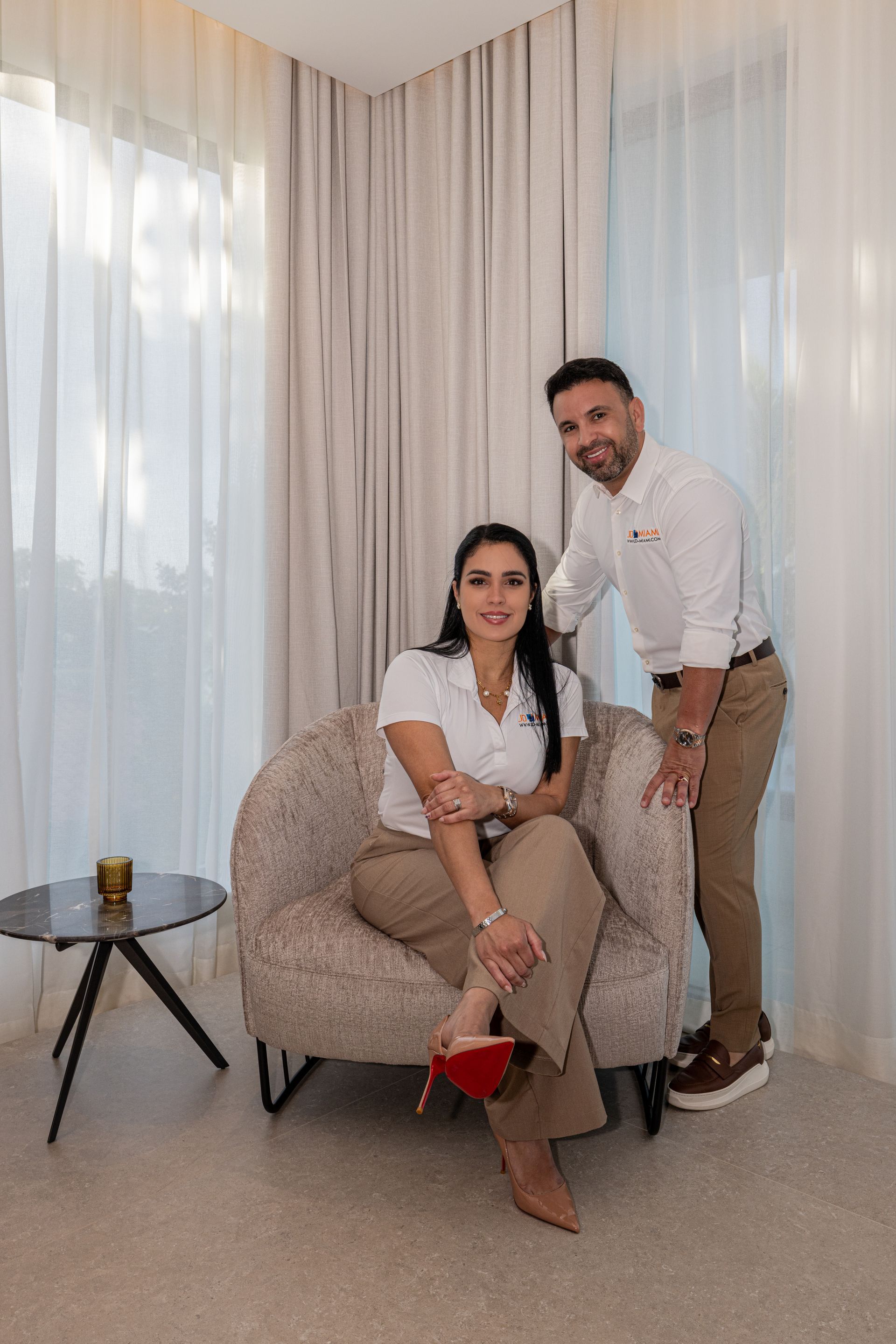 Couple in neutral-toned room. Woman sits in chair, man stands beside. They smile at camera near windows with sheer curtains.