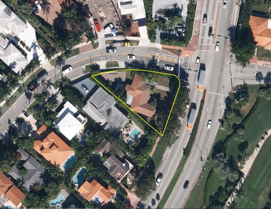 Aerial view of a house outlined in yellow, near a road with traffic, surrounded by trees and other buildings.