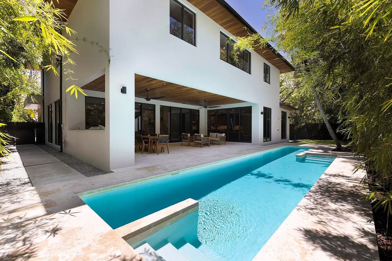 Two-story white house with a long, narrow pool. Outdoor patio with seating. Blue water, sunny day.