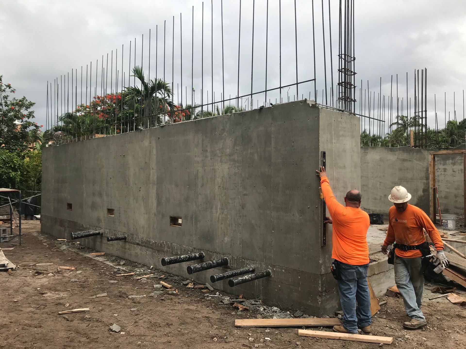 Two construction workers checking a concrete wall with rebar, outdoors, overcast.