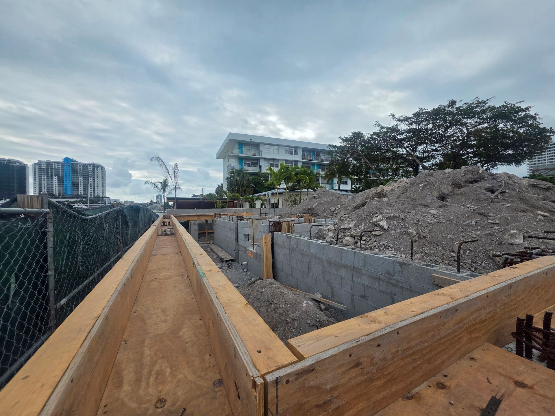 Construction site with wooden forms, concrete blocks, debris pile, buildings, and overcast sky.