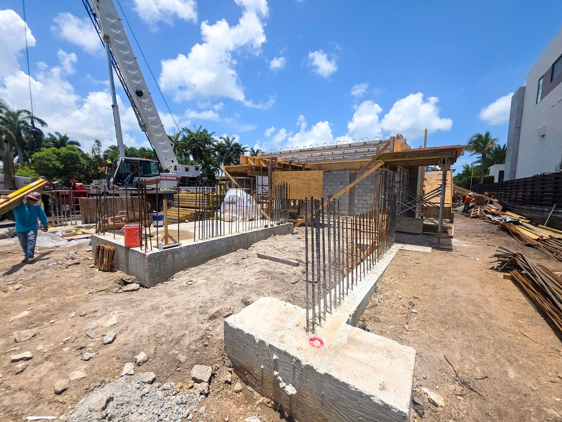 Construction site with exposed concrete foundations, rebar, and wooden forms. A crane and workers are present.