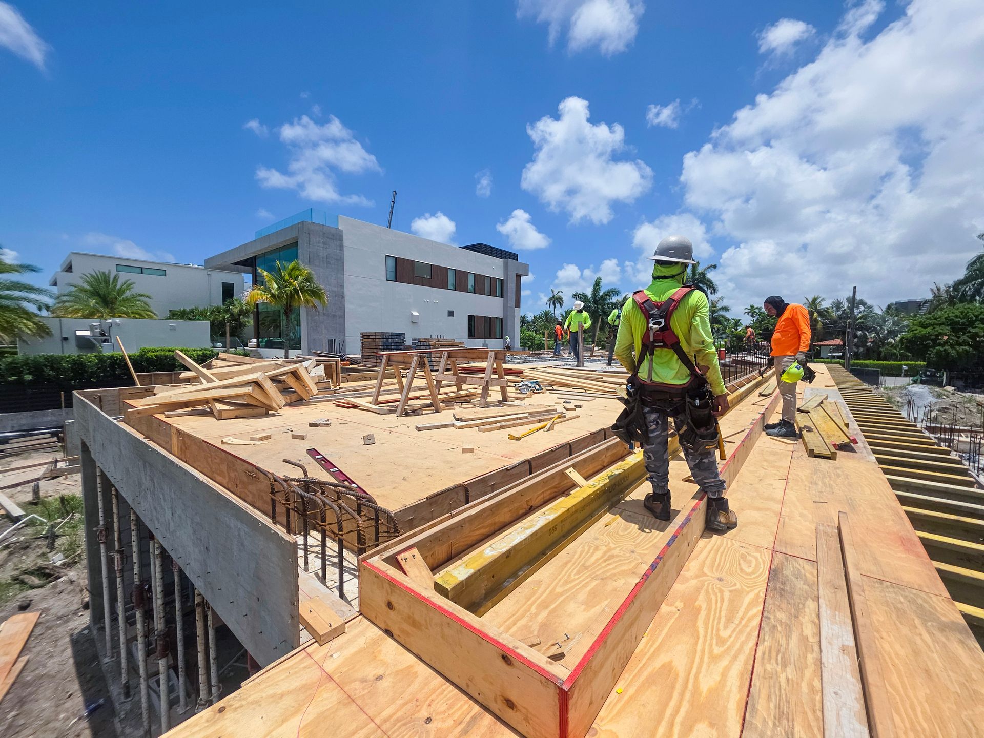 Construction workers on a rooftop, wearing safety gear, working on wooden framework. Bright sunny day, modern building in background.
