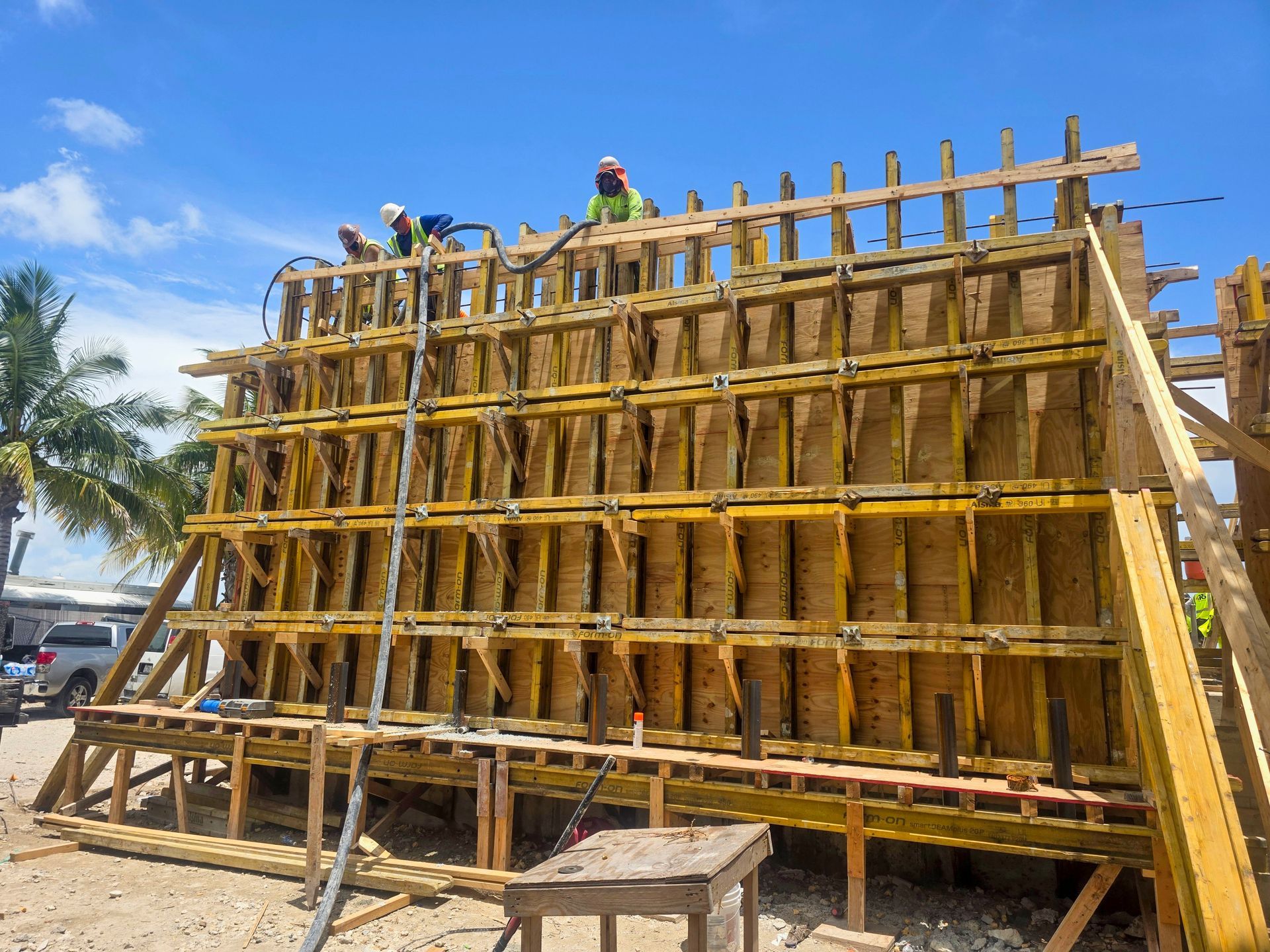 Construction workers on scaffolding, building a tall wooden formwork structure outdoors under a blue sky.