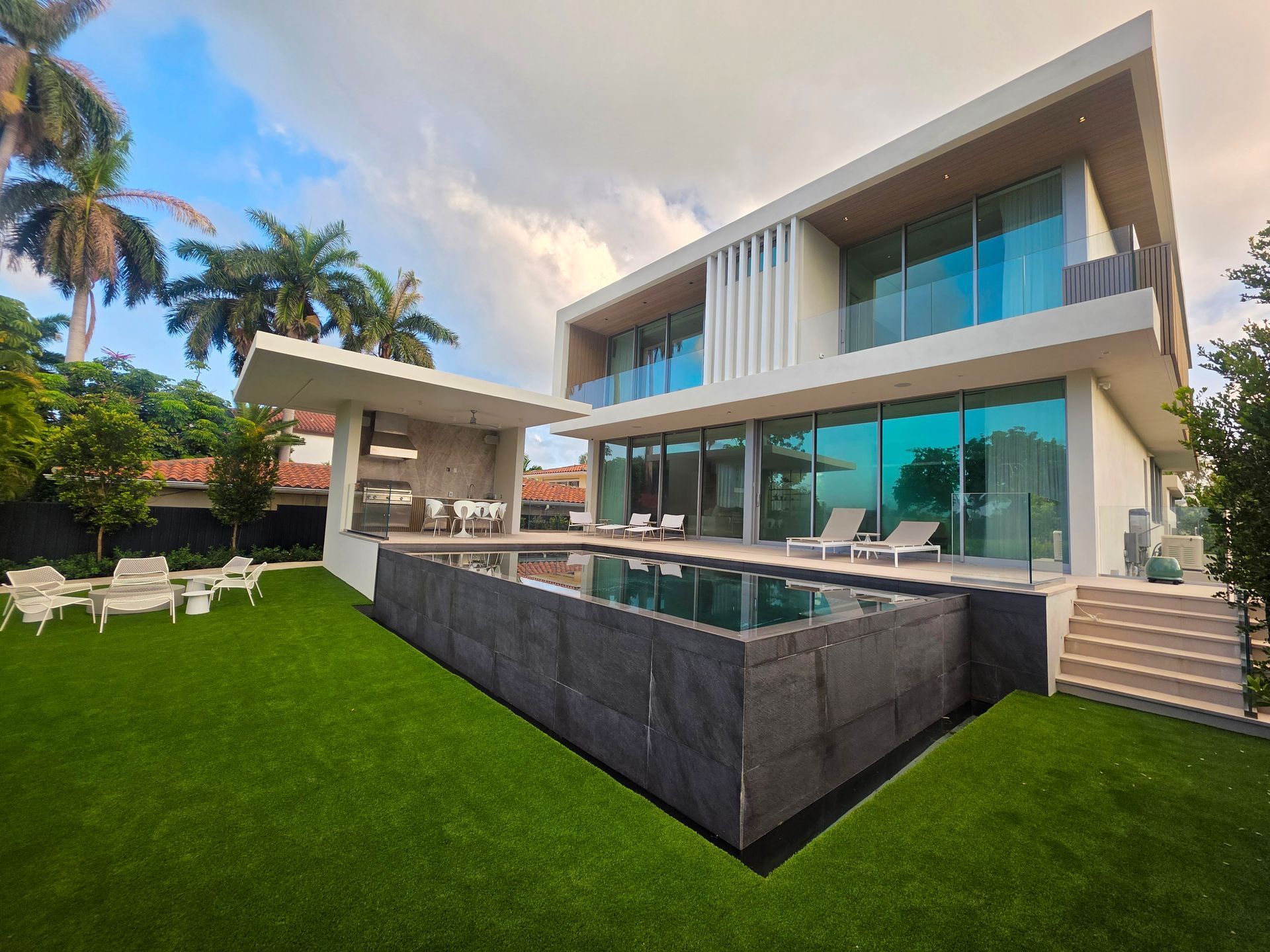 Modern two-story house with a pool and outdoor lounge area on a green lawn; cloudy sky.