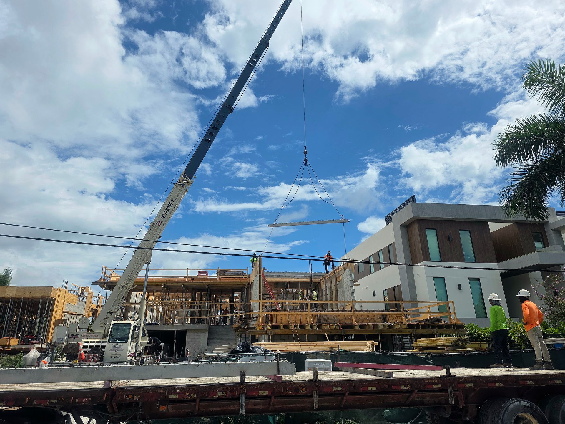 Construction site with crane lifting wooden frame near a multi-story building. Cloudy blue sky.