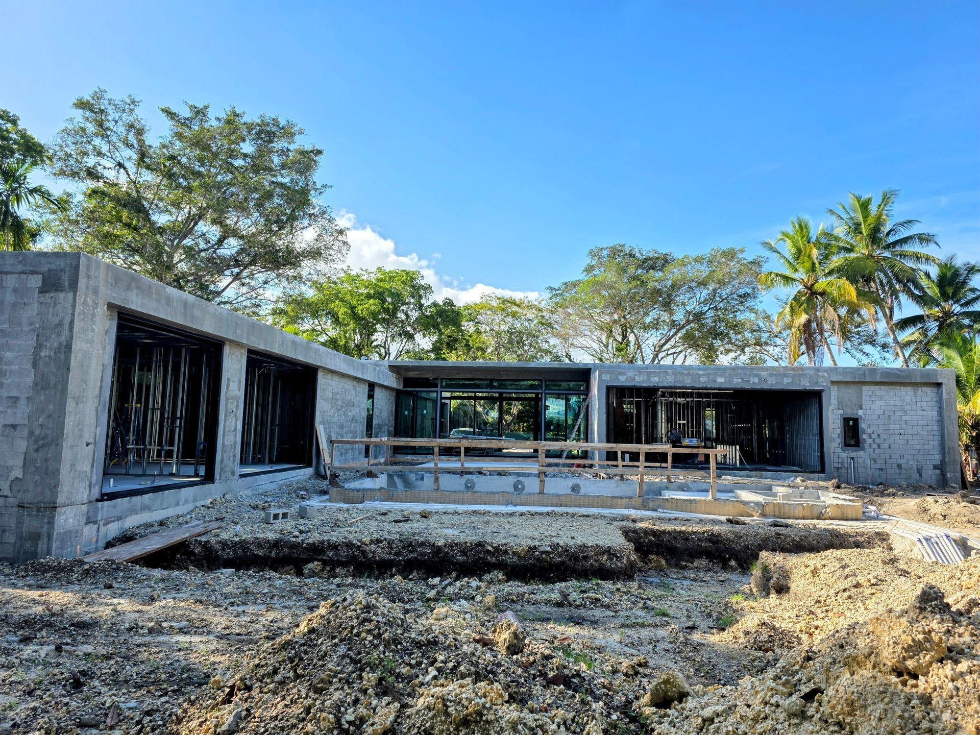 Construction site: modern concrete home under construction with surrounding trees and blue sky.